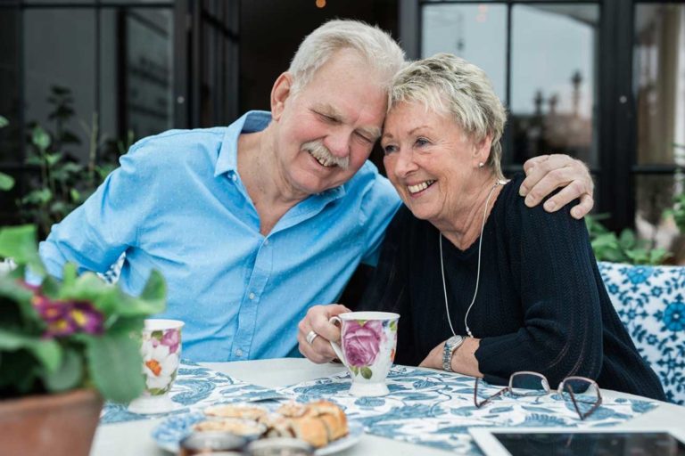 Äldre par som fikar med blommiga muggar och bullar och håller om varandra