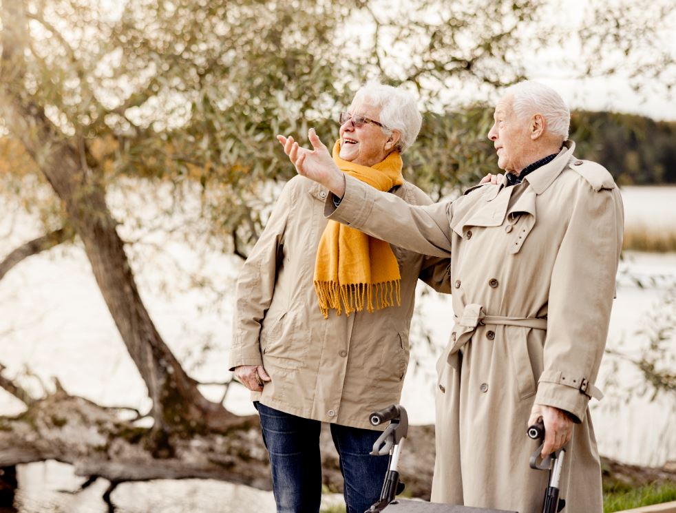 Äldre par som står vid en sjö och träd och ser glada ut